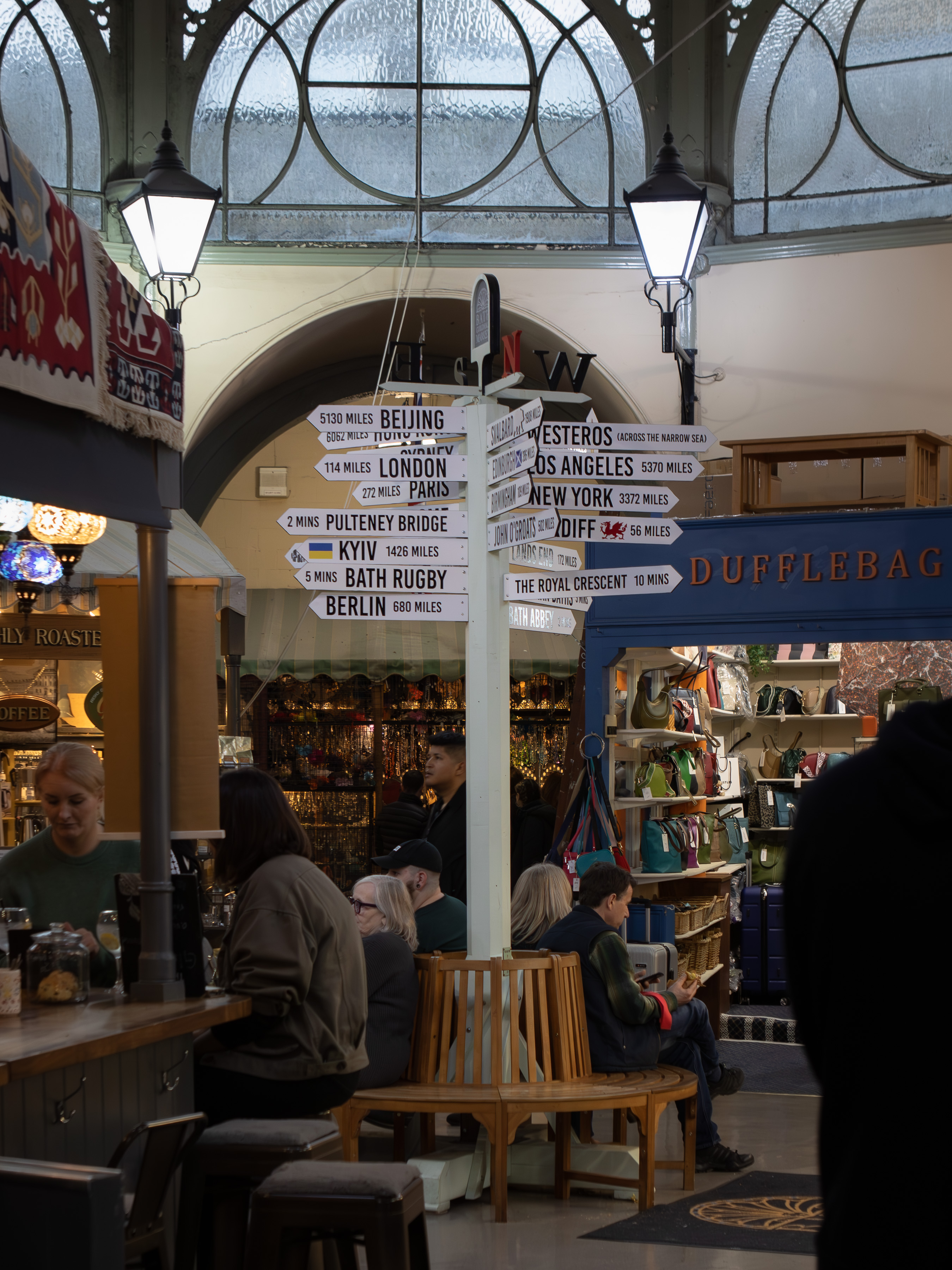 Bath signpost beneath the Victorian glass roof of the covered market pointing to Pulteney Bridge, London and Westeros, with covered market stalls beyond